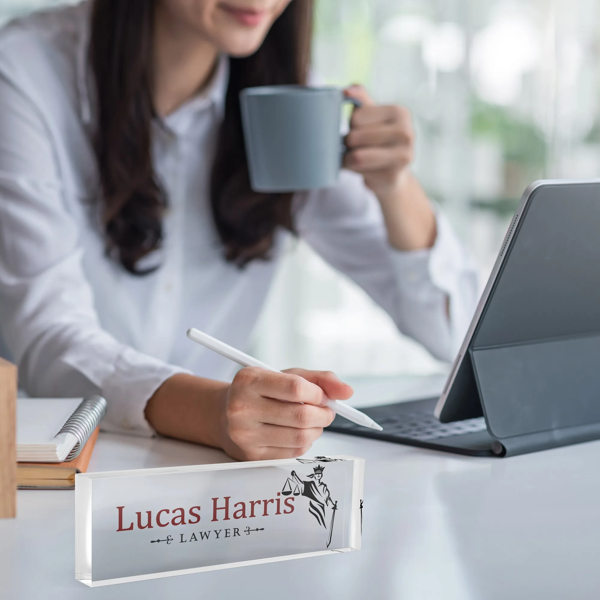Acrylic desk name plate with a Lady Justice silhouette design on a white desk beside a tablet and a coffee mug