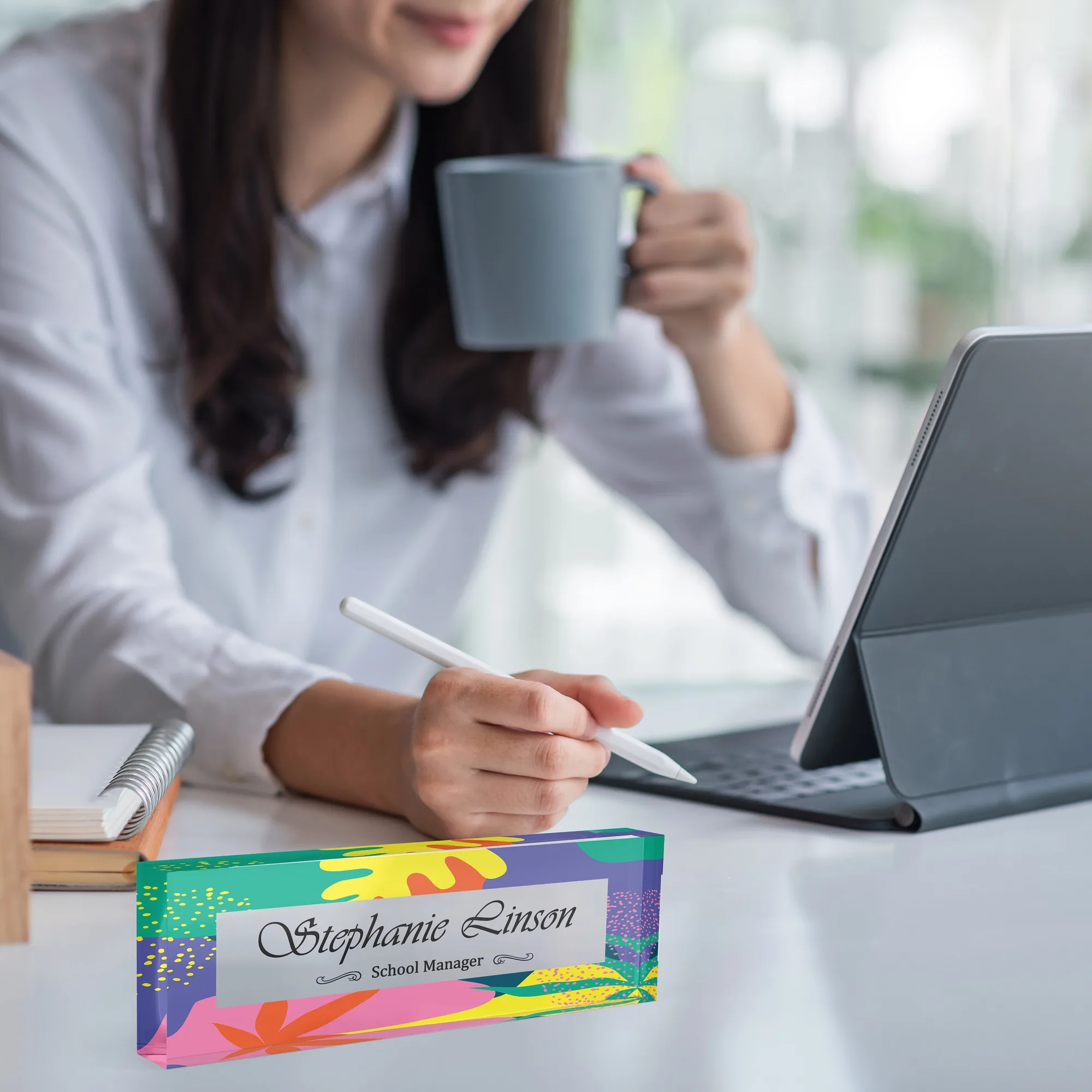 Acrylic desk name plate with a pink and yellow tropical design on a white desk beside a tablet and a coffee mug