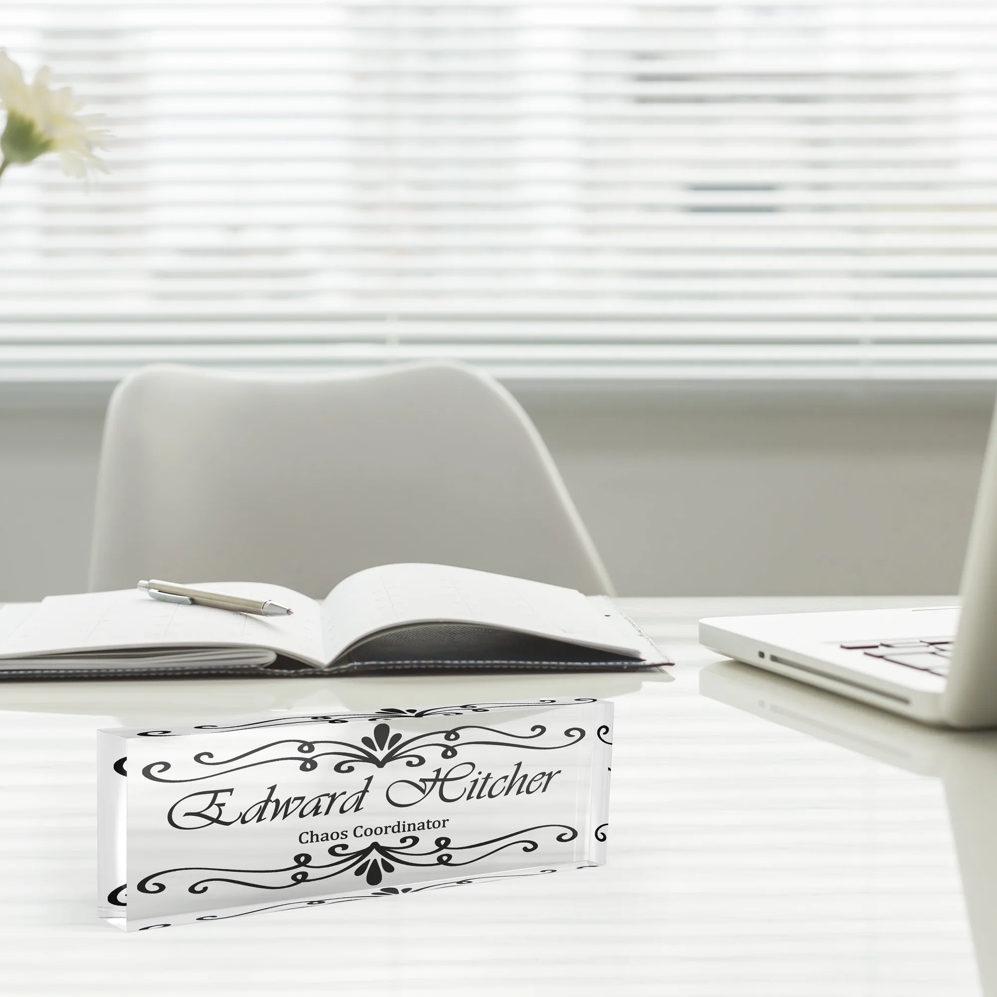 Acrylic desk name plate with a black Victorian scroll design beside an open notebook and a laptop