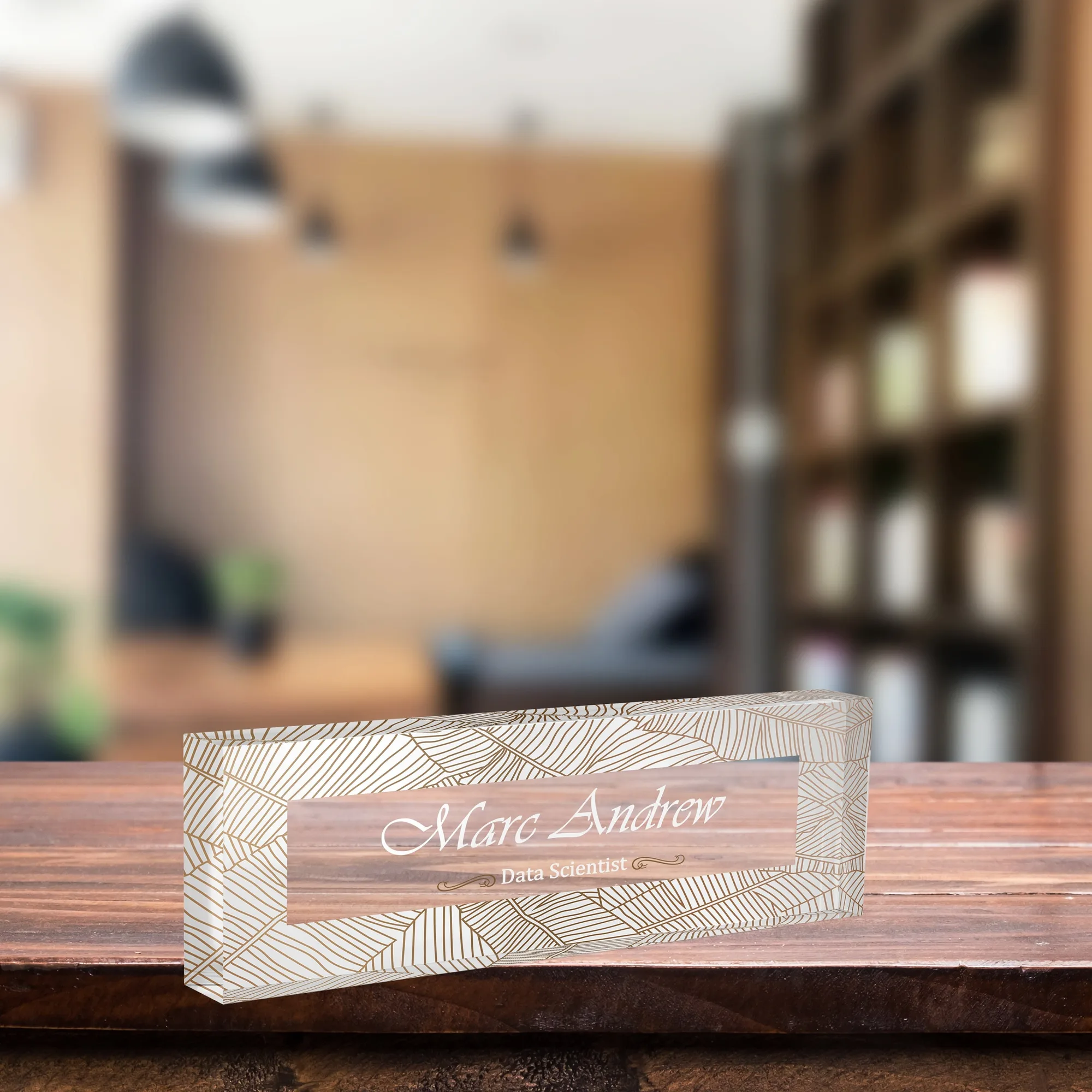 Clear acrylic name plate with a fine gold line leaf design on a wooden desk in a softly lit office